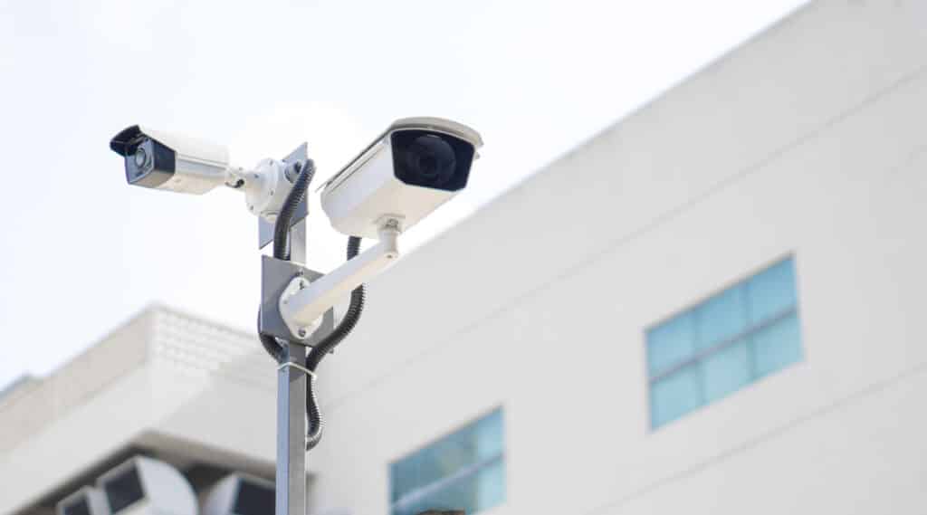 Caméras de surveillance extérieures fixes sur poteau blanc Deux caméras de surveillance blanches montées sur un poteau gris devant un bâtiment blanc, câbles noirs visibles.