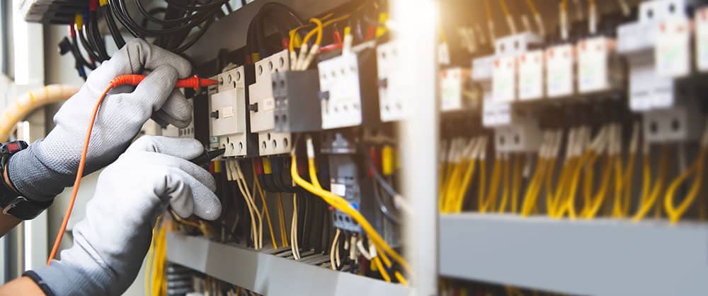 Maintenance Électrique : Test de Panneau Industriel Électricien portant des gants gris testant un panneau de contrôle électrique industriel avec un multimètre, câbles jaunes et composants visibles.