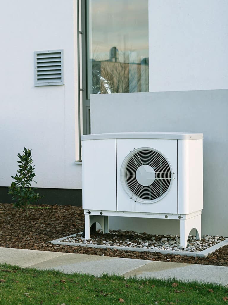 Pompe à Chaleur Air-Eau : Installation Moderne Unité extérieure de pompe à chaleur blanche, moderne, installée sur un socle près d'un mur blanc, avec une grille de ventilation et un petit arbre.