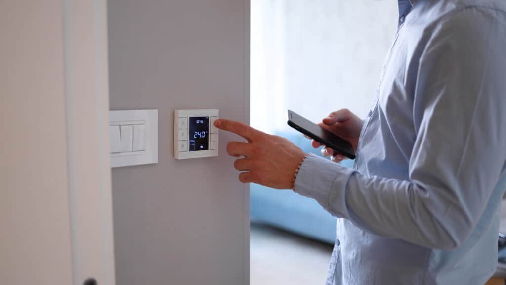 Thermostat intelligent 24.0°C : réglage et contrôle par smartphone Main ajustant un thermostat intelligent mural affichant 24.0°C, l'utilisateur tient un smartphone.